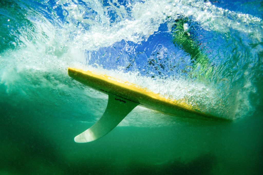 Underwater shot of yellow surfboard with single fin and a silhouette of female surfer above the water