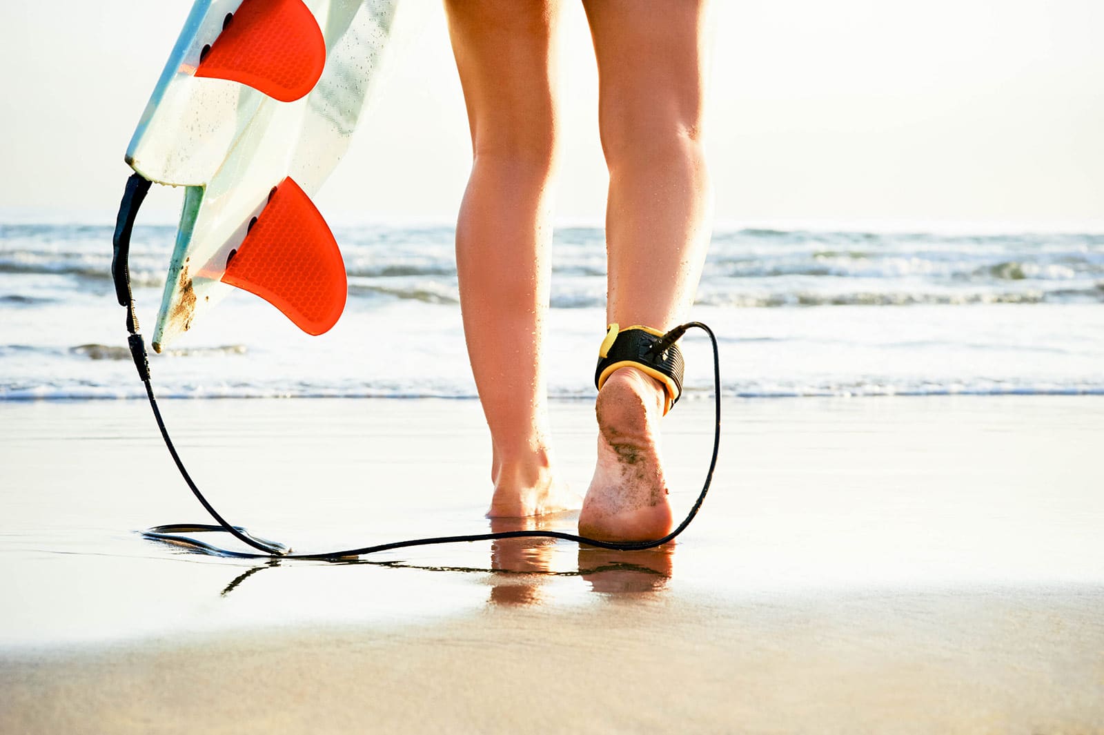 Close-up of woman's bare legs walking toward the ocean with a surfboard leash attached to her ankle