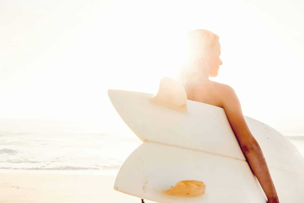Female surfer holding a fish surfboard under her arm with a dreamy sun flare behind her